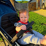 Kai is registered to the contest to win money with this photo: baby, stroller, child, outdoor, sunlight, grass, fence, sidewalk, car, tree, residential, red_clothing, blue_stroller, happy, person, infant, seatbelt, playful, summer, portrait