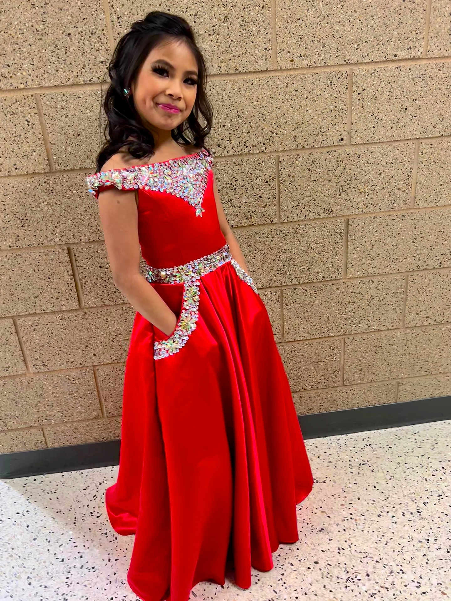 Avyannah is registered to the contest to win money with this photo: child, girl, red_dress, formal_dress, sparkles, embellishment, long_hair, makeup, smile, pose, brick_wall, hallway, tile_floor, pocket, portrait, elegant, pageant, young_person, standing, fashion