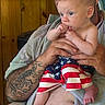 Zander is registered to the contest to win money with this photo: adult, american_flag, baby, child, closeup, curious, fingers, foot, hand, holding, indoor, infant, person, shirtless, shorts, sitting, skin, tattoo, towel, wooden_wall