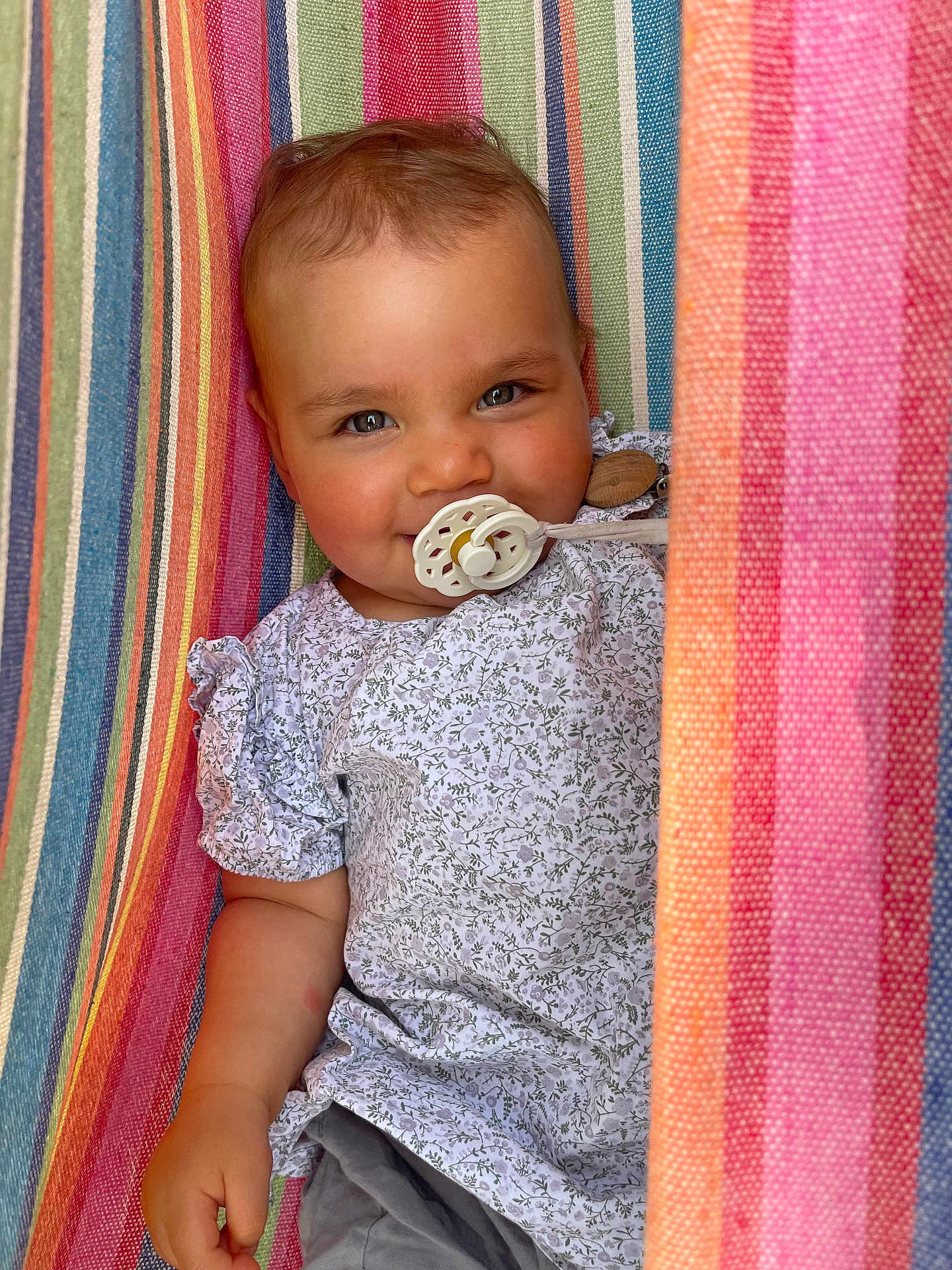 Adèle a rejoint le concours — aidez-le/la à gagner de superbes lots ! baby, pacifier, smiling, striped_hammock, colorful, floral_shirt, short_sleeves, child, cute, portrait, relaxed, outdoor, infant, happy, person, clothing, fabric, pattern, closeup, skin