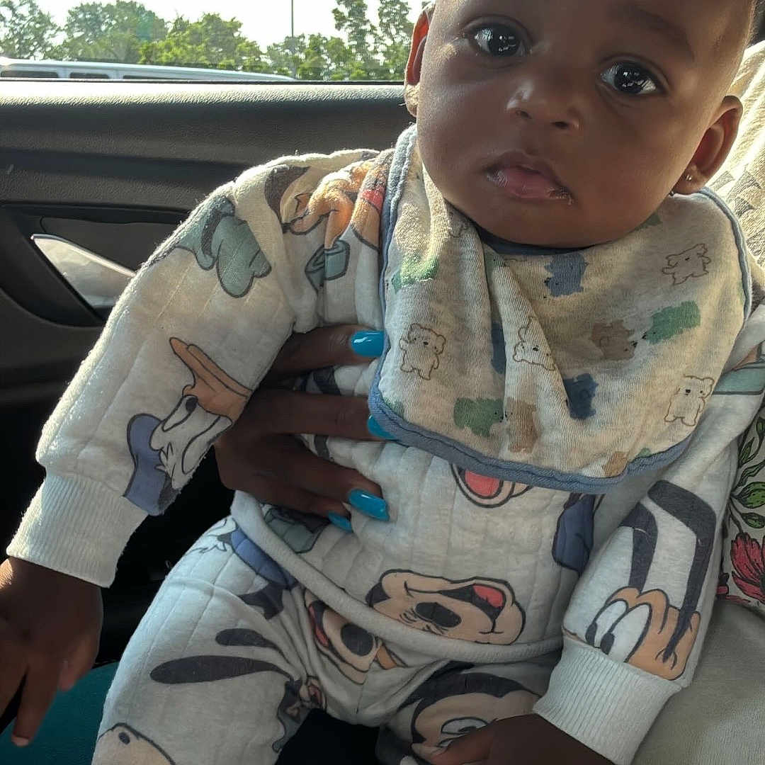 Iyon joined the competition — help win amazing prizes! baby, bib, blue_nail_polish, car_interior, cartoon_clothing, child, cozy_outfit, curious_look, dark_skin, daylight, hand, indoor, infant, light_pole, long_sleeves, pants, portrait, sitting, trees, window