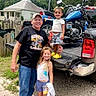 casual_clothing, child, children, daylight, family, fence, footwear, grass, house, jeans, man, motorcycle, nature, outdoor, pickup_truck, smiling, tailgate, tree, tshirt, vehicle