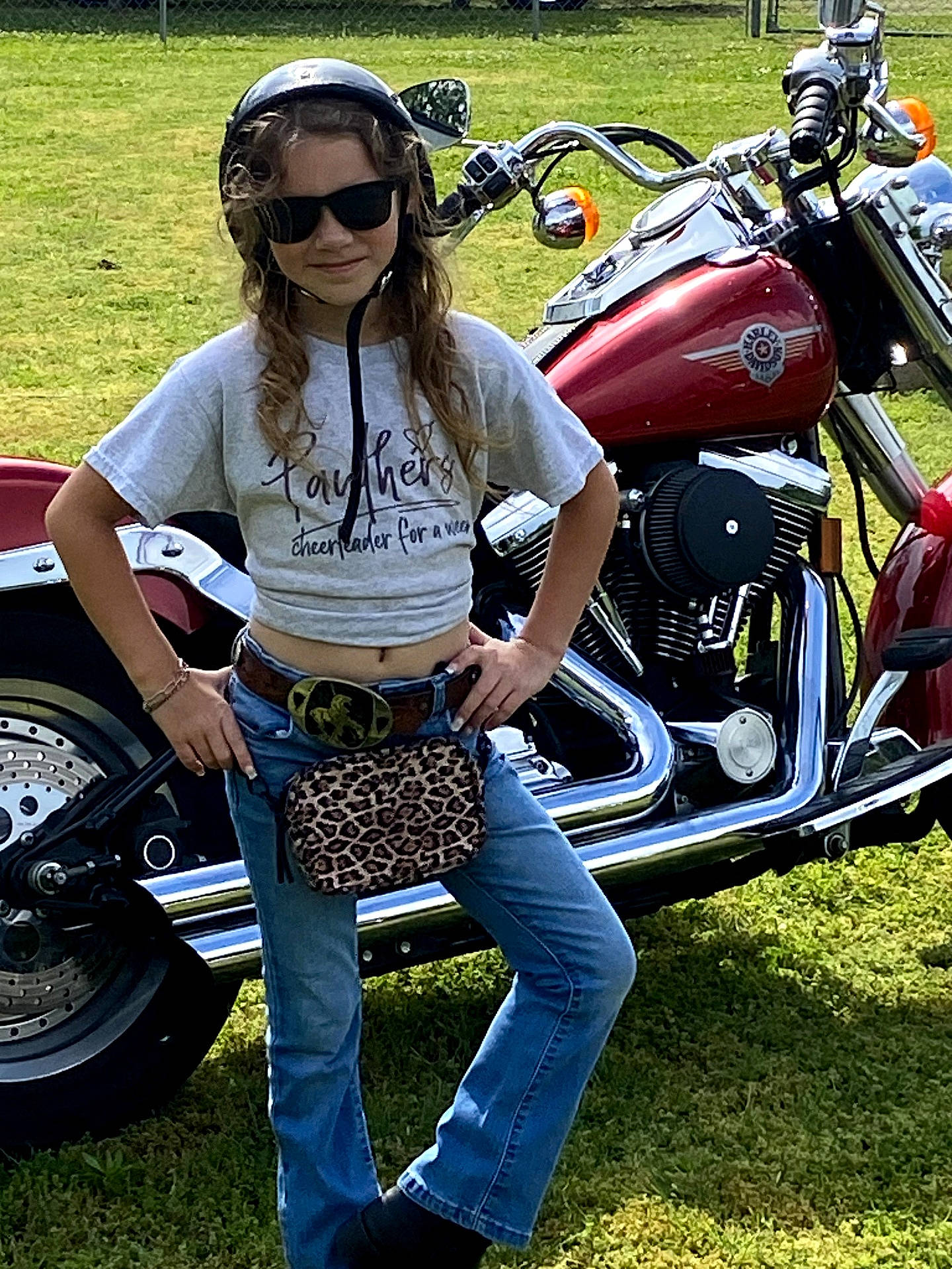 accessory, bag, belt, casual_clothing, child, chrome, girl, grass, helmet, jeans, leopard_print, metal, motorcycle, outdoor, pose, smile, summer, sunglasses, sunlight, vehicle