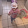 toddler, child, cap, white_cap, pink_toy, toy_vehicle, tiled_floor, kitchen, towels, playful, happy, motion_blur, indoor, person, smiling, clothing, leggings, socks, curly_hair, home