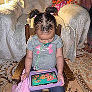 Cheyanne is registered to the contest to win money with this photo: armchair, blanket, cartoon, child, cozy, floor, girl, hair_buns, indoor, jeans, living_room, pacifier, pacifier_clip, rocking_chair, rug, sitting, sneakers, t_shirt, tablet, toddler