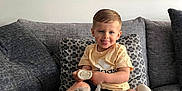 Meyson a rejoint le concours — aidez-le/la à gagner de superbes lots ! toddler, child, couch, sofa, plush_toys, pacifier, socks, cushions, indoor, smiling, tongue_out, short_hair, yellow_shirt, black_shorts, painting, wall_art, living_room, happy, person, home