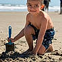 child, boy, smile, beach, sand, shovel, blue_shovel, swim_trunks, barefoot, ocean, waves, sunny, playing, sandcastle, portrait, outdoors, holiday, family, brunette, cute
