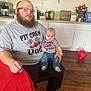 man, baby, tshirt, glasses, beard, kitchen, table, chair, balloon, floor, cabinet, bottle, flower, bag, person, indoor, family, casual, jeans, pacifier