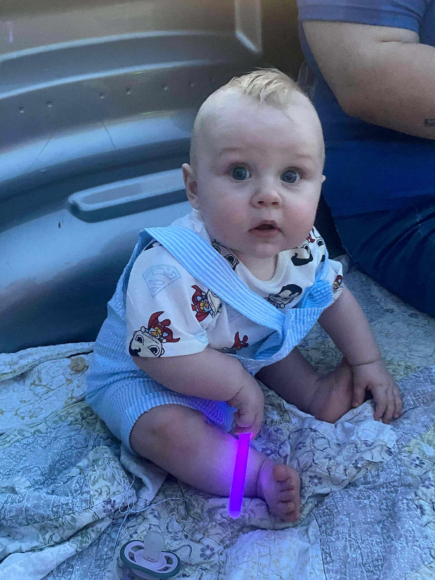 Cayden joined the competition — help win amazing prizes! baby, child, infant, sitting, blue_overalls, superhero_tshirt, pacifier, glowing_stick, purple_light, quilt, blanket, indoor, person, cute, curly_hair, wide_eyes, hand, foot, adult_arm, casual_clothing