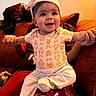 baby, child, headband, smiling, person, couch, cushion, orange, red, clothing, sitting, happy, indoors, portrait, infant, hand, holding, cute, cozy, warm_lighting