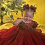 child, toddler, girl, red_dress, bow, holiday, christmas_tree, ornament, lights, smile, happy, indoor, portrait, curly_hair, sitting, plush_rug, festive, sparkle, wall, photography