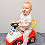 toddler, child, toy_car, steering_wheel, smiling, indoor, light_hair, blue_eyes, white_polo_shirt, beige_pants, sock, red_toy, yellow_handle, gray_blanket, playful, cute, baby, person, sitting, portrait