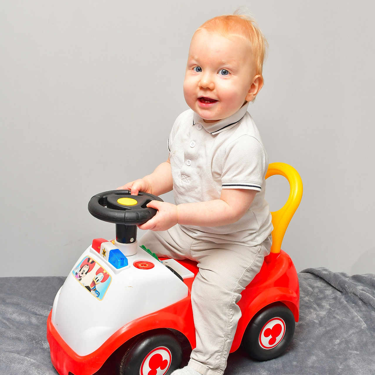 Ayden participe au concours pour gagner de l'argent avec cette photo : baby, beige_pants, blue_eyes, child, cute, gray_blanket, indoor, light_hair, person, playful, portrait, red_toy, sitting, smiling, sock, steering_wheel, toddler, toy_car, white_polo_shirt, yellow_handle