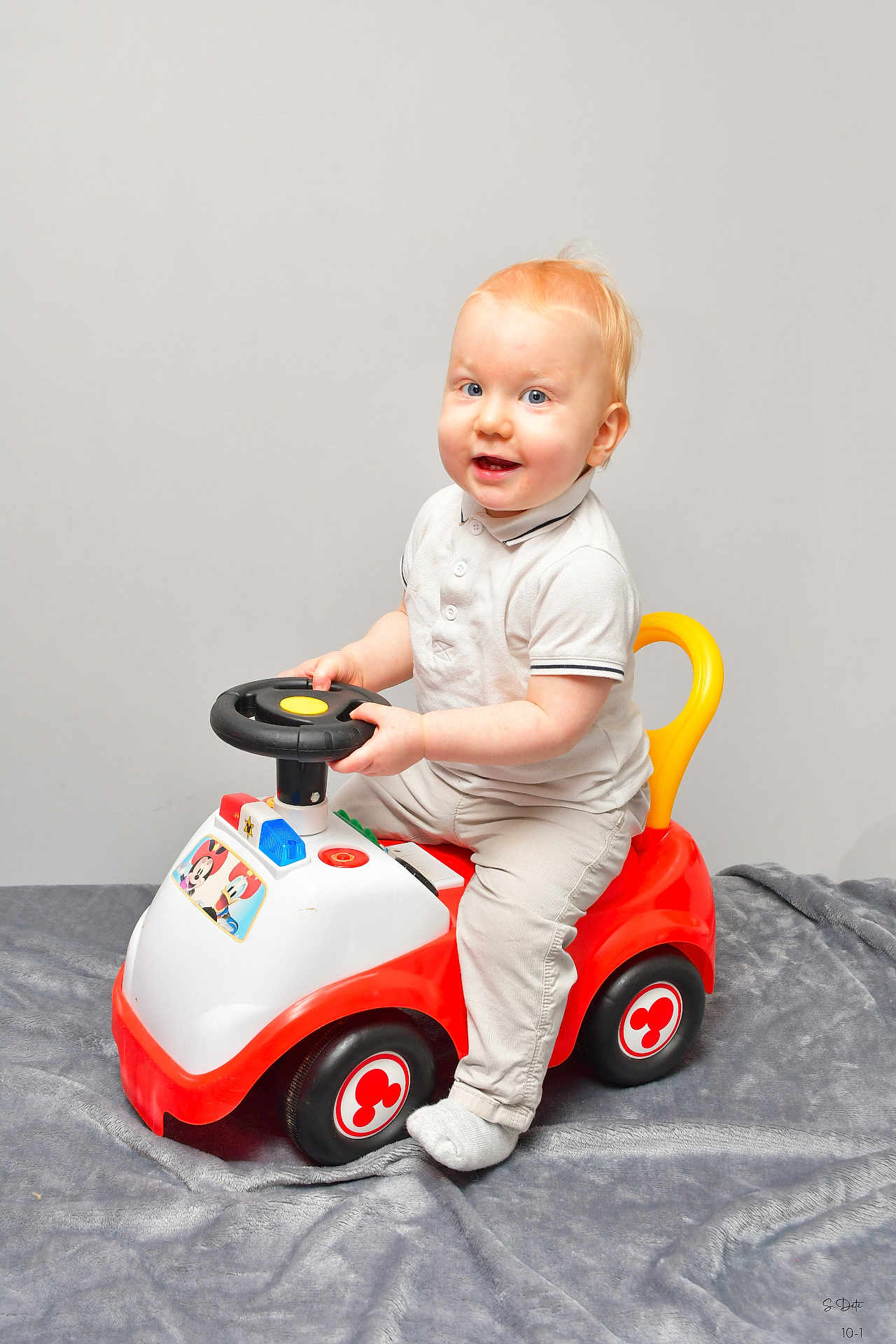 Ayden participe au concours pour gagner de l'argent avec cette photo : toddler, child, toy_car, steering_wheel, smiling, indoor, light_hair, blue_eyes, white_polo_shirt, beige_pants, sock, red_toy, yellow_handle, gray_blanket, playful, cute, baby, person, sitting, portrait