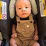 baby, infant, car_seat, overalls, clothing, seat_belt, safety_warning, warning_label, head_support, face, legs, feet, hands, person, child, indoor, cute, portrait, newborn, seat