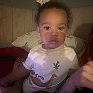 Amarii is registered to the contest to win money with this photo: toddler, child, indoor, portrait, white_shirt, dinosaur_graphic, pillow, wood_paneling, curly_hair, tattoo, adult_arm, casual_clothing, person, face, seated, looking_at_camera, home, soft_lighting, expression, baby