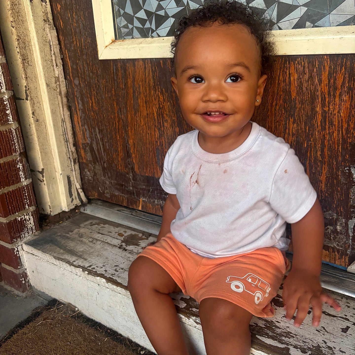Amarii joined the competition — help win amazing prizes! barefoot, brick_wall, child, curly_hair, cute, daylight, doorstep, earring, happy, orange_shorts, outdoor, patterned_glass, playful, portrait, sitting, smiling, toddler, white_tshirt, window, wooden_door