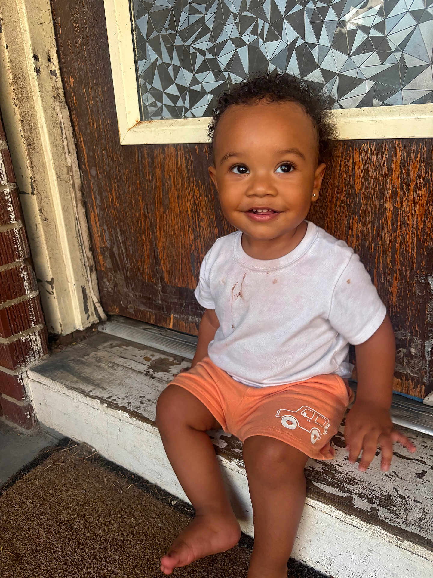 Amarii joined the competition — help win amazing prizes! child, toddler, smiling, curly_hair, earring, white_tshirt, orange_shorts, barefoot, doorstep, wooden_door, brick_wall, outdoor, window, patterned_glass, happy, sitting, portrait, cute, playful, daylight