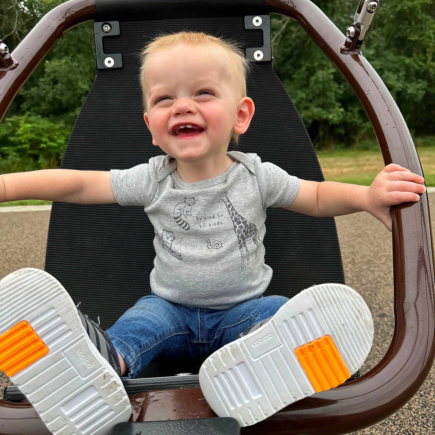 Everitt is registered to the contest to win money with this photo: animal_print, arms_outstretched, child, daylight, fun, greenery, happy, jeans, joy, nature, outdoor, park, play, playground, seat, short_sleeve_shirt, smiling, sneakers, swing, toddler