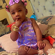 Royalty is registered to the contest to win money with this photo: toddler, child, baby, indoor, bed, pillow, blanket, purple_outfit, unicorn_pattern, hair_bows, bracelets, necklace, curious_expression, sitting, cozy, bedroom, soft_lighting, cute, person, portrait