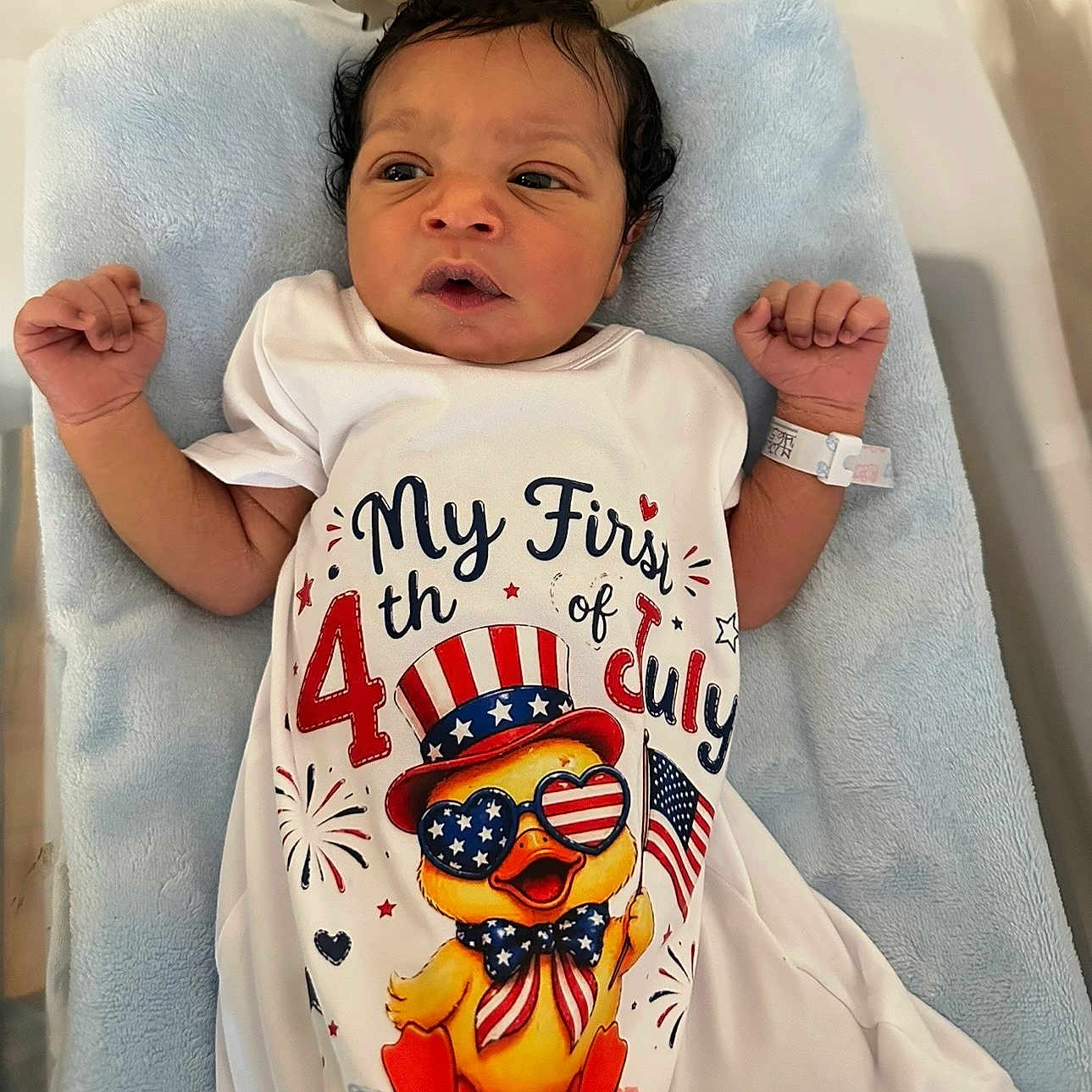 Khazari is registered to the contest to win money with this photo: 4th_of_july, baby, blue_blanket, celebration, clothing, cute, duck_illustration, expression, face, holiday, hospital_bracelet, indoor, infant, lying_down, newborn, onesie, patriotic, person, portrait, small_hands