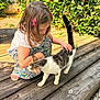child, cat, petting, outdoor, wooden_deck, greenery, sunlight, tabby_cat, girl, summer, nature, cute, animal, young_child, casual_clothing, hair_accessory, barefoot, daytime, playful, affection