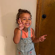 Octavia is registered to the contest to win money with this photo: toddler, child, smiling, peace_sign, curly_hair, denim_dress, orange_shirt, indoor, wooden_door, wall, happy, portrait, person, cute, gesture, standing, playful, young_child, fashion, casual