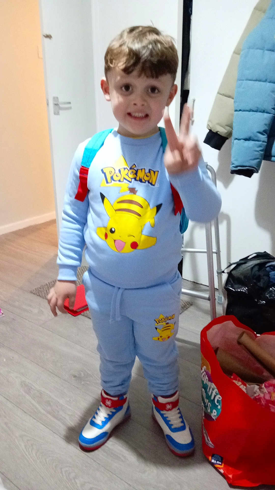 Grayson Francis is registered to the contest to win money with this photo: child, boy, indoor, pokemon, clothing, backpack, peace_sign, smiling, standing, shoes, floor, door, coat, bag, toy, person, casual, happy, young, room