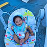 baby, child, smiling, curly_hair, baby_rocker, colorful, animal_print, outdoor, blue_floor, yellow_stripes, steps, happy, infant, seat, playful, footwearless, toddler, person, portrait, seatbelt