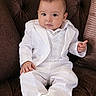 armchair, baby, big_eyes, brown_upholstery, child, cute, formal_clothing, indoor, infant, lap, looking_at_camera, photography, portrait, short_hair, sitting, small_hands, socks, tufted_chair, vest, white_suit