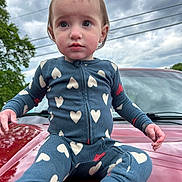 Harlynn is registered to the contest to win money with this photo: child, baby, onesie, heart_pattern, car, hood, red, outdoor, cloudy_sky, tree, curious, portrait, sitting, clothing, infant, person, casual, expression, daylight, closeup