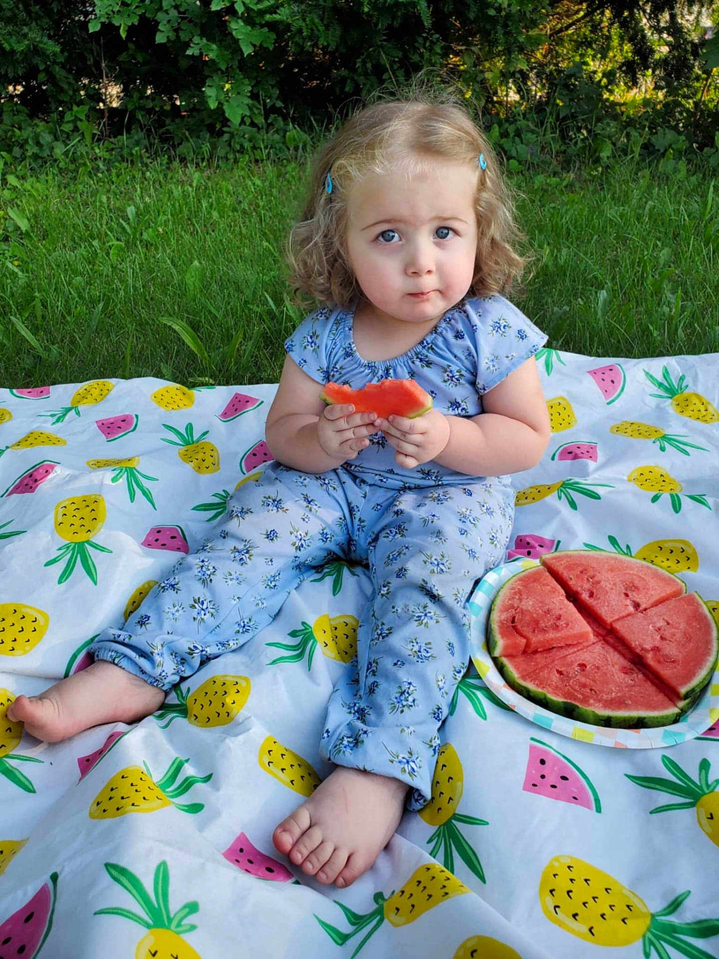 Addison joined the competition — help win amazing prizes! toddler, child, watermelon, blanket, outdoor, grass, curly_hair, blue_eyes, sitting, fruit, summer, snack, plate, nature, cute, clothing, patterned_blanket, picnic, expression, barefoot