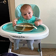 Mason is registered to the contest to win money with this photo: baby, child, high_chair, feeding, food, plate, spoon, bib, smiling, portrait, indoor, kitchen, seated, infant, turquoise, messy_eating, wood_floor, door, wall, family_photo