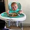baby, child, high_chair, feeding, food, plate, spoon, bib, smiling, portrait, indoor, kitchen, seated, infant, turquoise, messy_eating, wood_floor, door, wall, family_photo