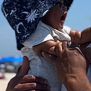 Zaydan is registered to the contest to win money with this photo: baby, child, hat, sun_hat, adult_hand, beach, outdoor, sky, summer, holding, close_up, person, infant, happy, daylight, casual_clothing, skin, portrait, nature, vacation