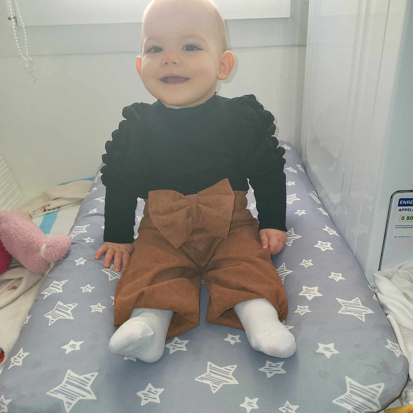 Slyvia a rejoint le concours — aidez-le/la à gagner de superbes lots ! baby, black_top, bow, brown_pants, changing_pad, child, clothing, cozy, cute, furniture, happy, indoor, infant, light, person, seated, smiling, socks, star_pattern, window