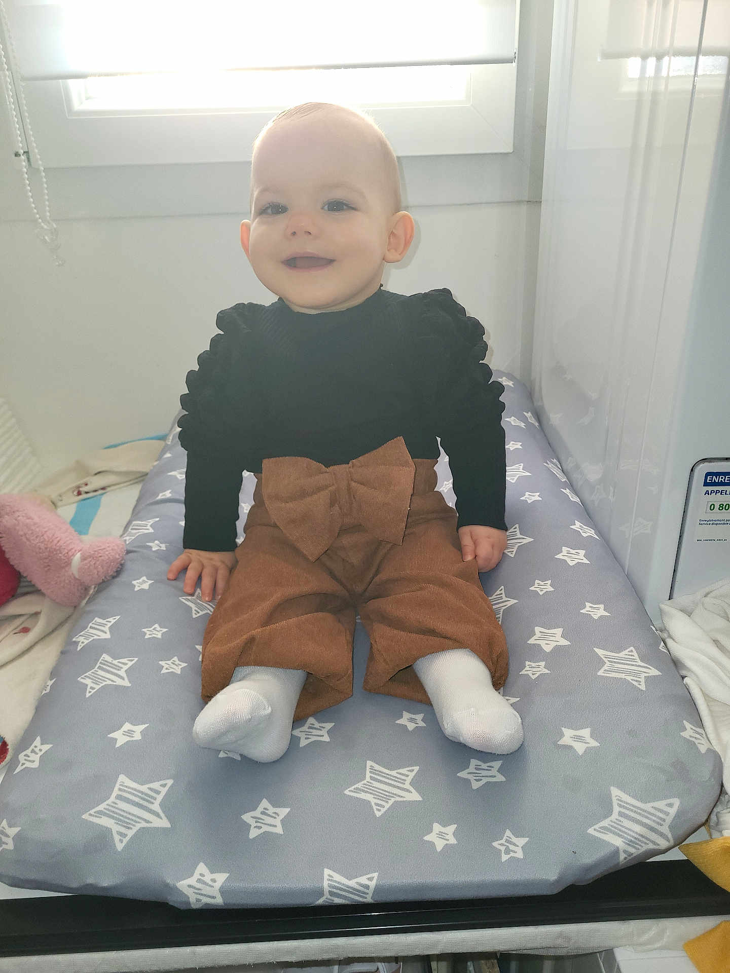Slyvia a rejoint le concours — aidez-le/la à gagner de superbes lots ! baby, black_top, bow, brown_pants, changing_pad, child, clothing, cozy, cute, furniture, happy, indoor, infant, light, person, seated, smiling, socks, star_pattern, window