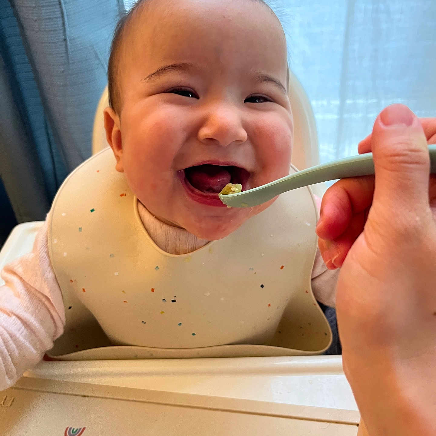 Yoona is registered to the contest to win money with this photo: baby, child, feeding, high_chair, bib, spoon, smiling, happy, indoor, person, face, hand, curtain, light, cute, infant, expression, clothing, table, meal