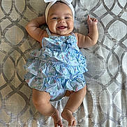 Zoe is registered to the contest to win money with this photo: baby, smiling, headband, bow, dress, patterned_background, happy, infant, cute, child, lying_down, blue, white, fabric, skin, cheeks, hands, legs, toes, indoors