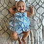 baby, smiling, headband, bow, dress, patterned_background, happy, infant, cute, child, lying_down, blue, white, fabric, skin, cheeks, hands, legs, toes, indoors