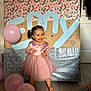 toddler, child, pink_dress, balloons, barefoot, smiling, happy, indoor, flooring, decorations, party, celebration, portrait, face, person, hair_bows, background, signage, holiday, festive