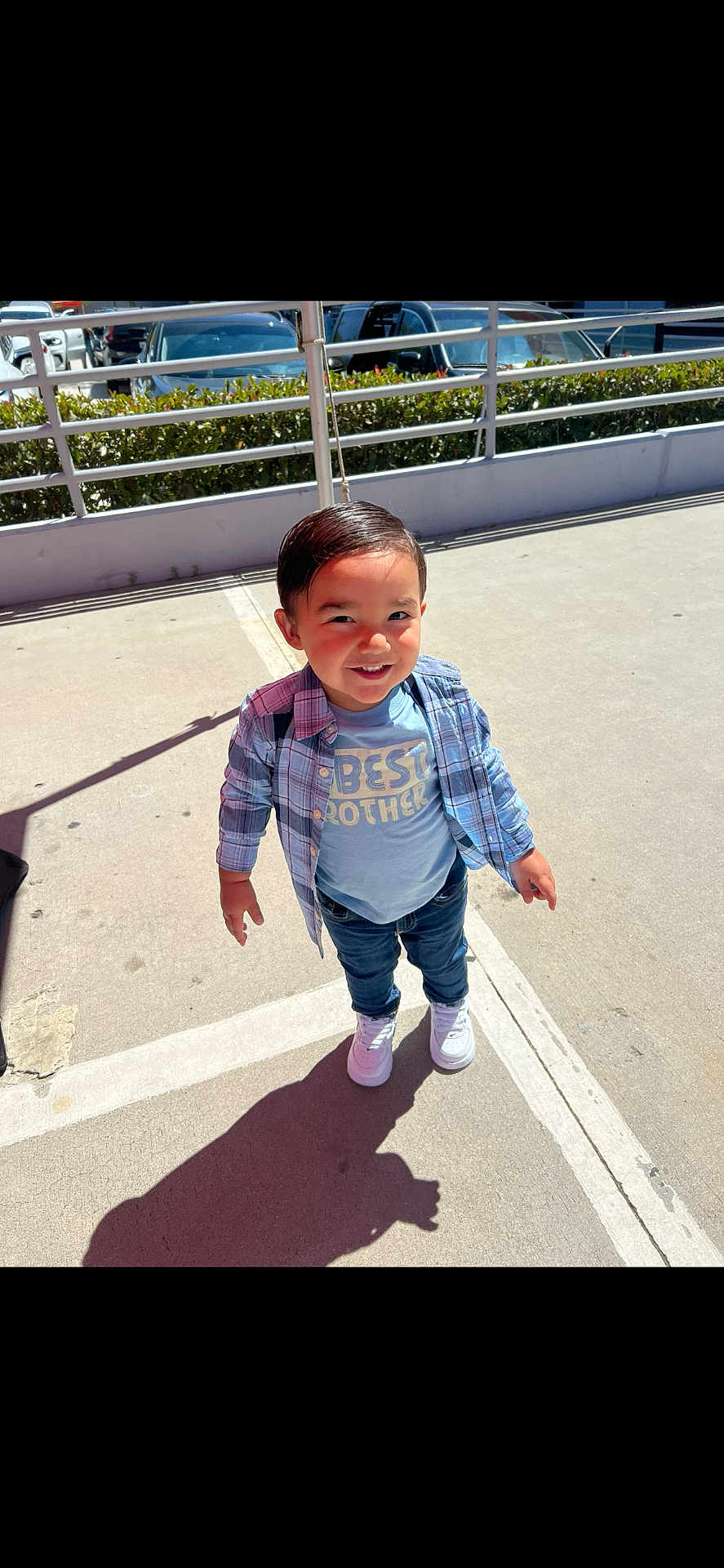 Carlos is registered to the contest to win money with this photo: child, toddler, boy, outdoor, sunny, plaid_shirt, blue_tshirt, best_brother, jeans, white_sneakers, smiling, shadow, concrete, parking_lot, railing, plants, daylight, casual_clothing, happy, standing