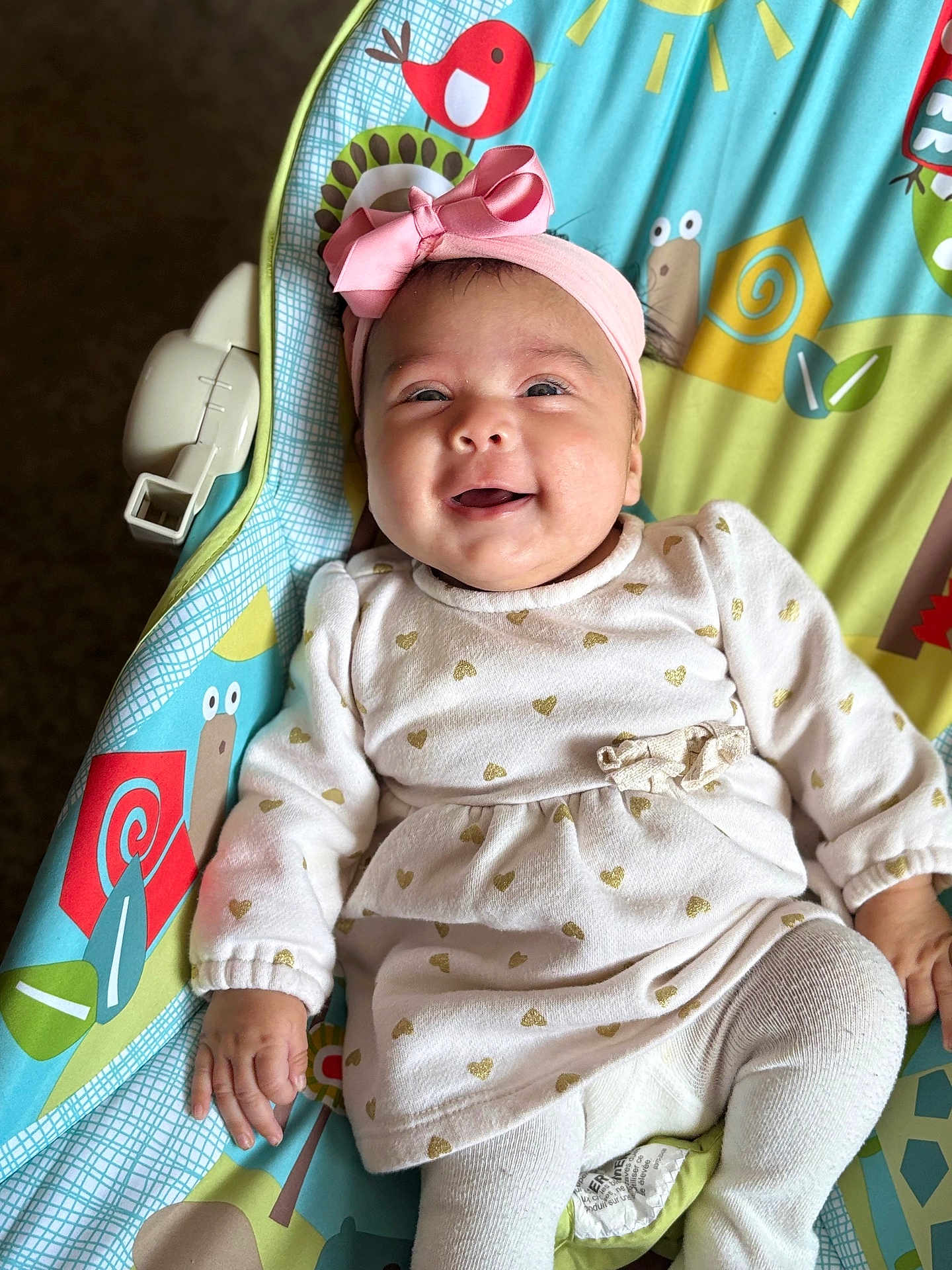 Merry is registered to the contest to win money with this photo: baby, baby_seat, bird, child, clothing, colorful, cute, gold_hearts, happy, headband, indoors, infant, laying_down, patterned, pink_bow, portrait, smiling, snail, toddler, white_dress