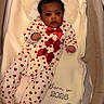 baby, infant, onesie, heart_pattern, red_bows, bassinet, blanket, pillow, cozy, indoor, sleeping_area, cute, portrait, newborn, baby_clothes, fabric, soft_texture, floor, wood, person