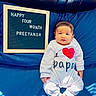 baby, infant, child, onesie, bed, blue_bedspread, letter_board, text, celebration, milestone, happy, cute, indoors, light_and_shadow, wood_panel, smiling, face, feet, person, clothing
