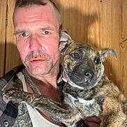 Mercy is registered to the contest to win money with this photo: adult_male, brindle, close_up, collar, dog, eyes, facial_hair, indoor, man, necklace, nose, paw, person, pet, plaid_shirt, portrait, puppy, selfie, tank_top, wood_paneling