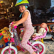 Lucy is registered to the contest to win money with this photo: child, helmet, bicycle, training_wheels, doll, toy, indoor, living_room, carpet, television, furniture, books, smile, pink, pants, shirt, sock, handlebars, helmet_strap, seat