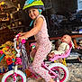 child, helmet, bicycle, training_wheels, doll, toy, indoor, living_room, carpet, television, furniture, books, smile, pink, pants, shirt, sock, handlebars, helmet_strap, seat