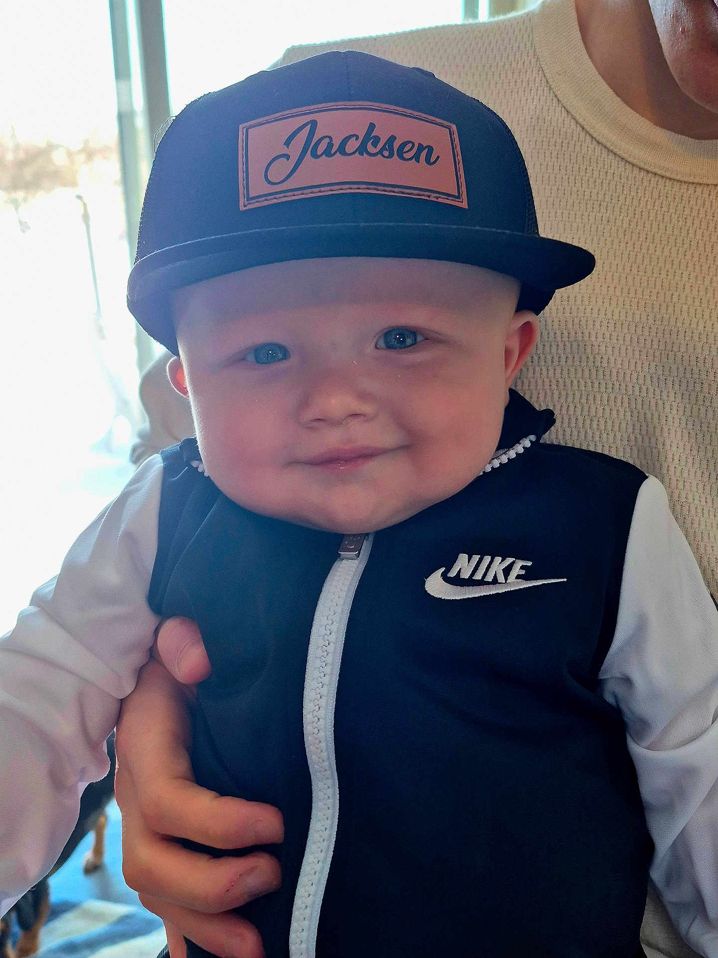 Jacksen is registered to the contest to win money with this photo: baby, infant, smile, cap, hat, jacket, nike_logo, face, blue_eyes, chubby_cheeks, portrait, indoor, hand, adult, holding, zipper, window_light, clothing, person, cute