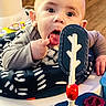 baby, child, tongue, hand, toy, activity_center, indoor, floor, curious, expression, play, infant, person, face, clothing, sitting, wood_floor, colorful, closeup, cute